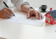 Título de propiedad y posesión: conceptos que debes conocer Persona firmando documento de escrituras junto a una maqueta de casa, representando la adquisición de un título de propiedad