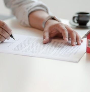 Título de propiedad y posesión: conceptos que debes conocer Persona firmando documento de escrituras junto a una maqueta de casa, representando la adquisición de un título de propiedad